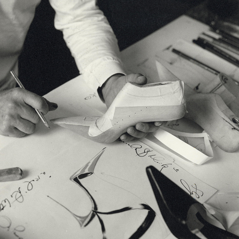 A close up of Manolo holding a shoe last. His other hand is resting on a table holding a pencil.