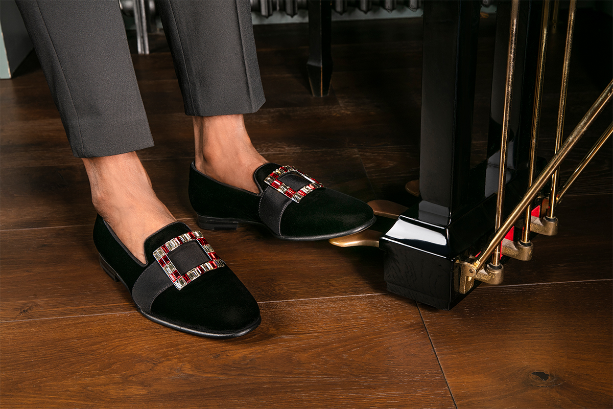 Model wearing velvet loafers with grosgrain ribbon and red and clear crystal buckle, playing the piano 