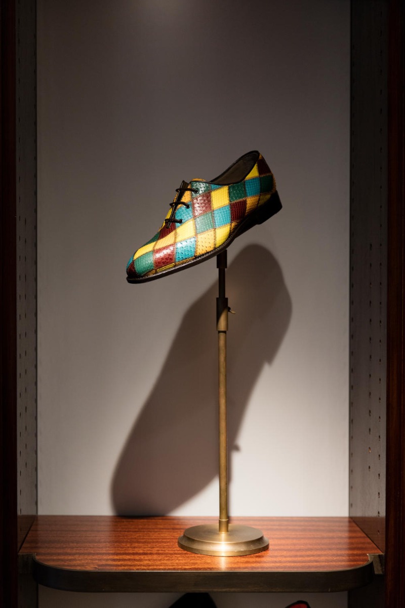A mens lace up shoe on a shoe stand. The shoe features yellow, blue, red and green diamonds in a harlequin pattern. 