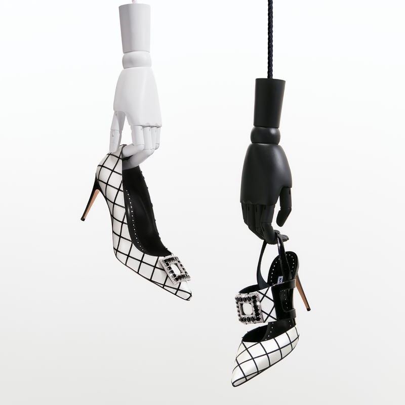 Wooden hands holding 2 black and white windowpane pumps with embellished detail.