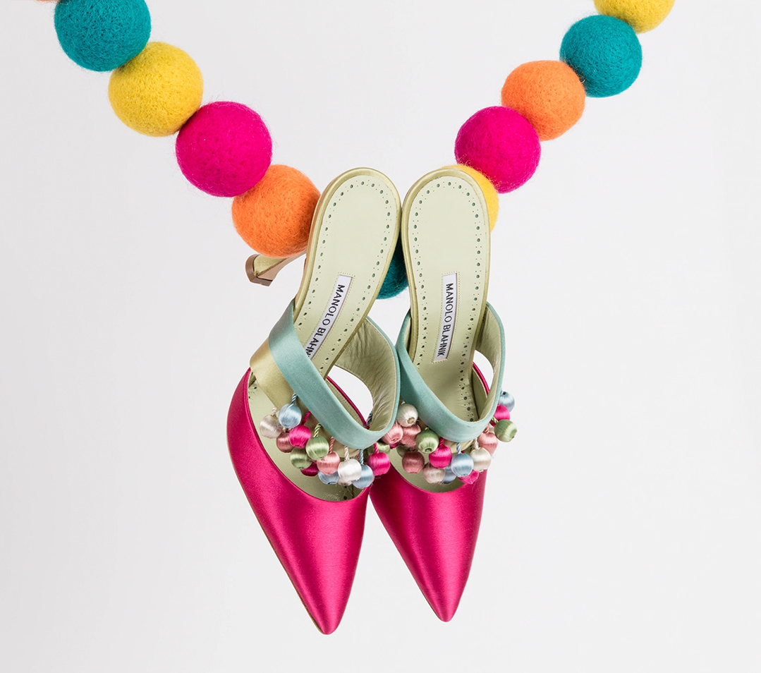 A pair of pink heeled mules with pom pom tassels. The mules are hanging from a chain of pom poms. 