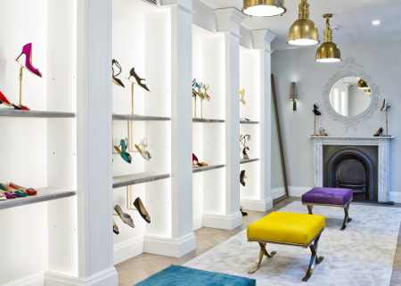 Upstairs in the Burlington Arcade store. On the left there are shelves with shoes. There are three stools in the centre. 