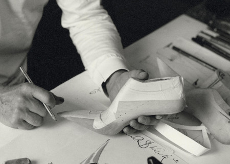 A close up of Manolo holding a shoe last. His other hand is resting on a table holding a pencil.