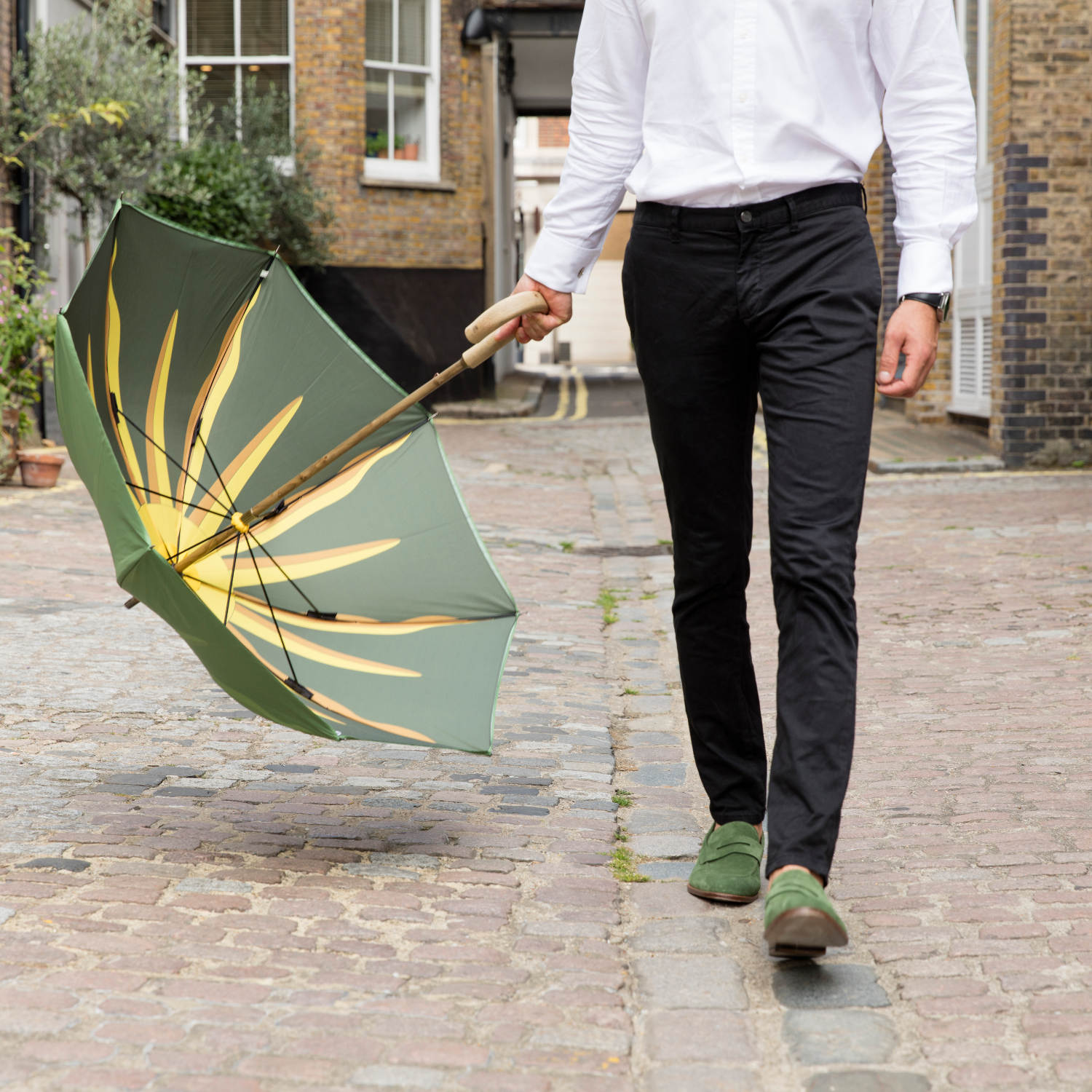 A man on a cobbled street wearing green loafers. He is holding a green umbrella with a sunburst print inside the umbrella.