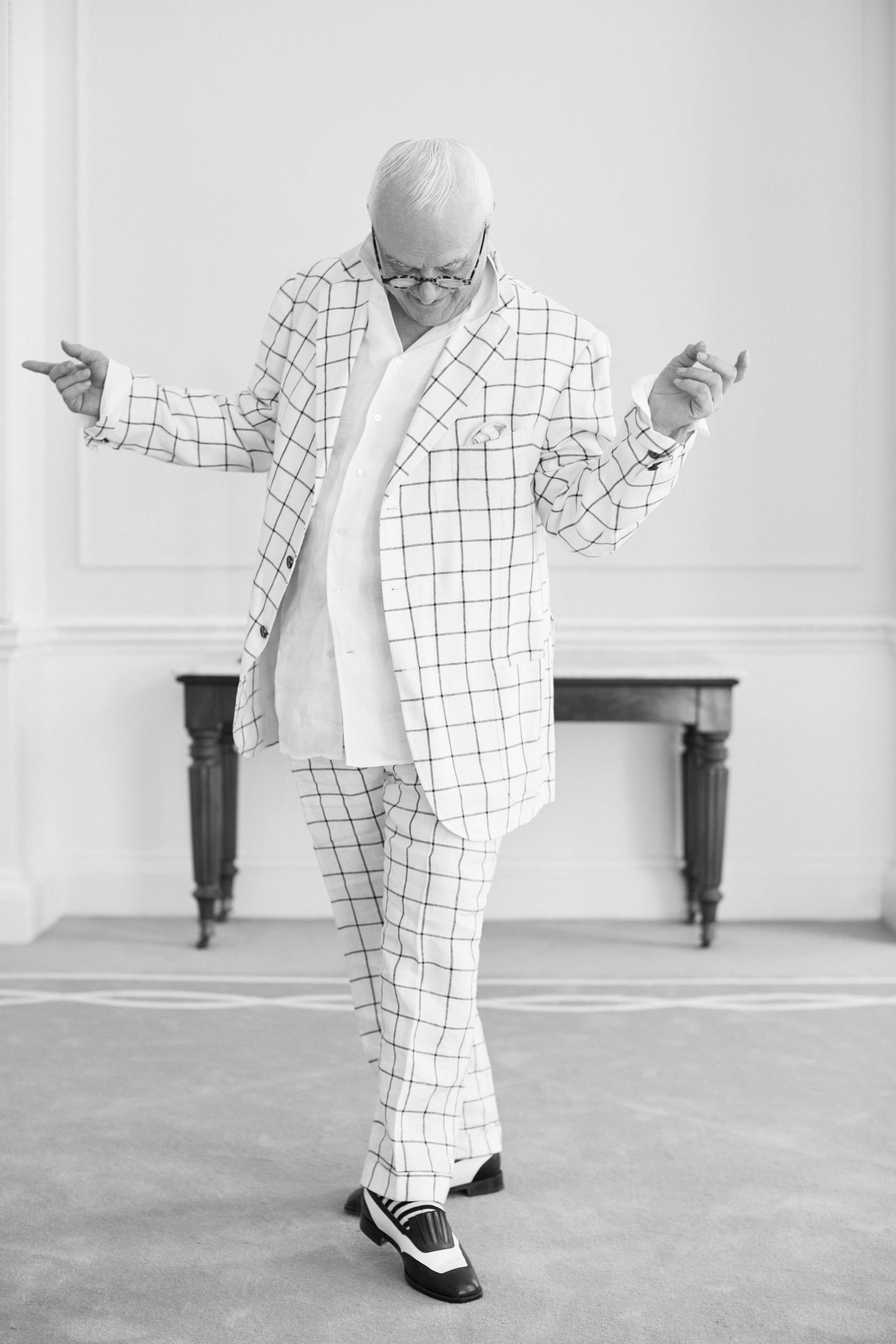Manolo Blahnik wearing a white suit. He is looking at his feet. His hands are up in a whimsical and fun position.