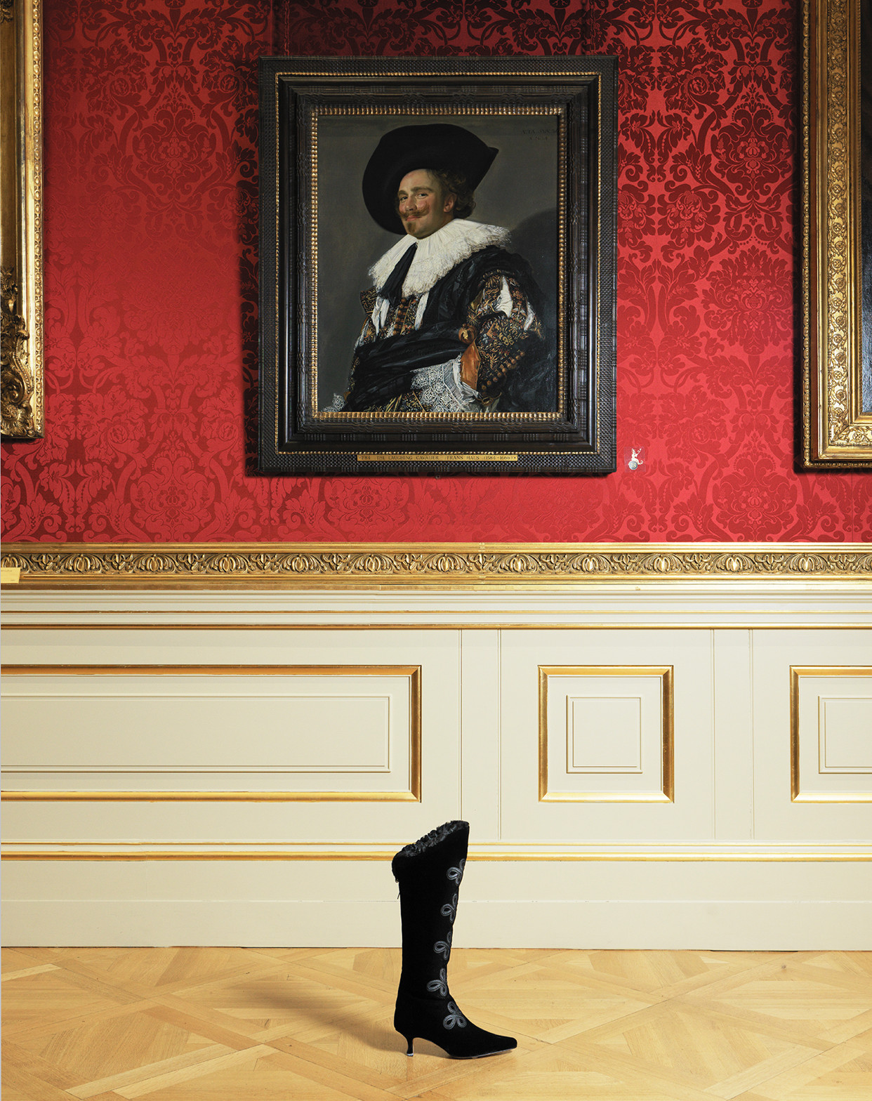 The Asha boot. A black knee high boot with ribbons, is positioned in front of The Laughing Cavalier painting by Frans Hals. 