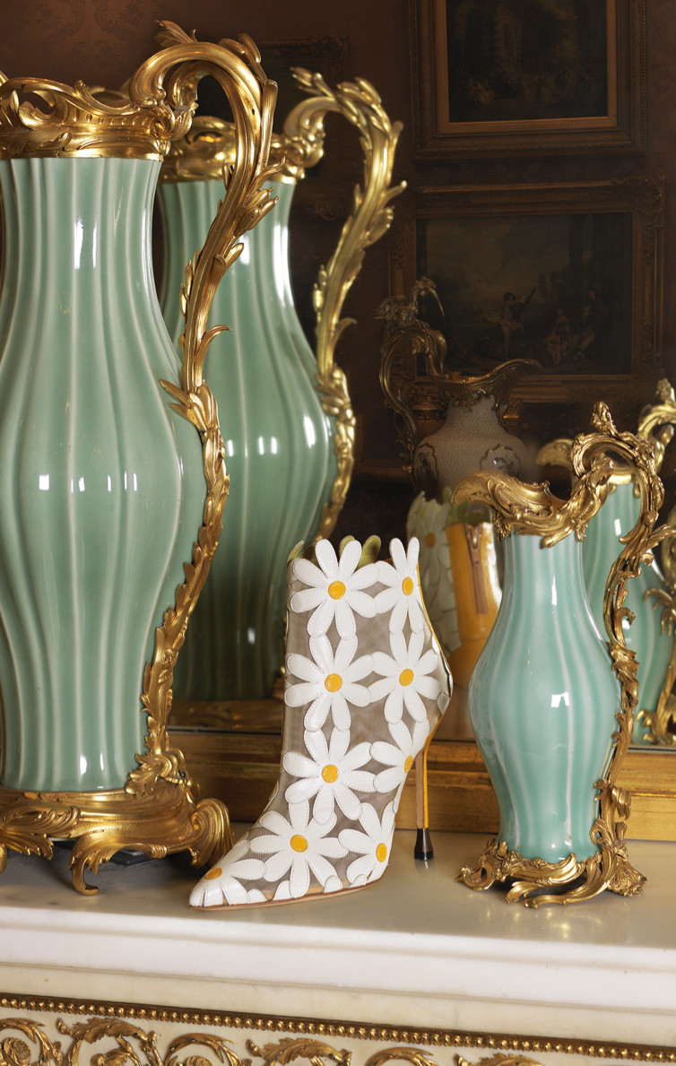 The Margolotta Boot. A boot featuring oversized daisies is positioned in front of a mirror and between two ornate vessels.