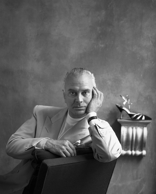 A black and white portrait of Manolo Blahnik. He is in the Old Church Street store, sitting on a chair.