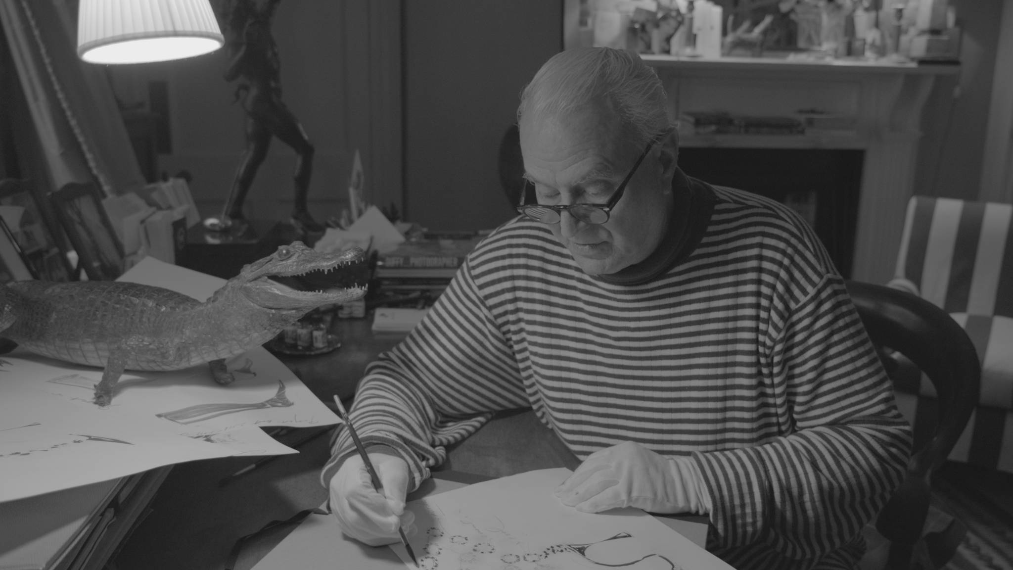 A black and white image of Manolo sketching a shoe. He is wearing a Breton turtle neck and white gloves.